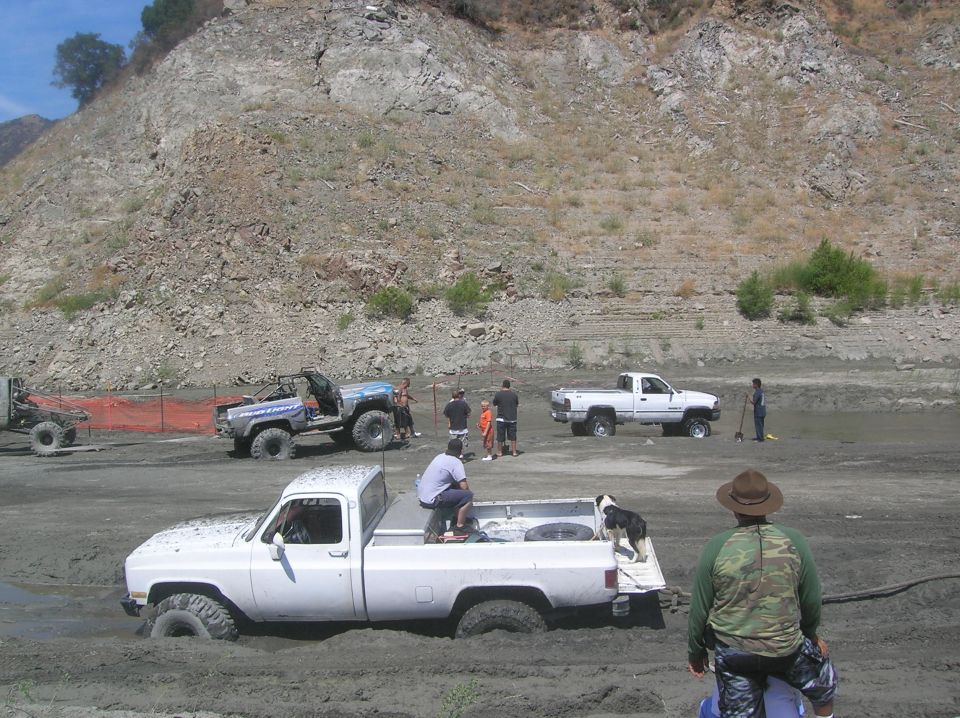 Several off-road vehicles are stuck in mud, with people around them. One white pickup truck is partially submerged