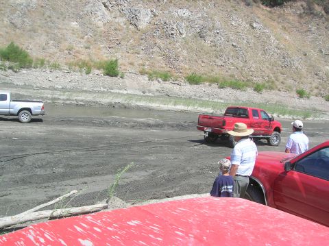 1985 Ford Bronco - Several people watch as a red pickup truck is stuck in the mud, with a silver truck in the background.