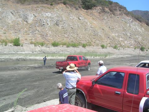 1985 Ford Bronco - Red pickup trucks are stuck in the mud near a dry riverbed with a rocky hillside in the background.