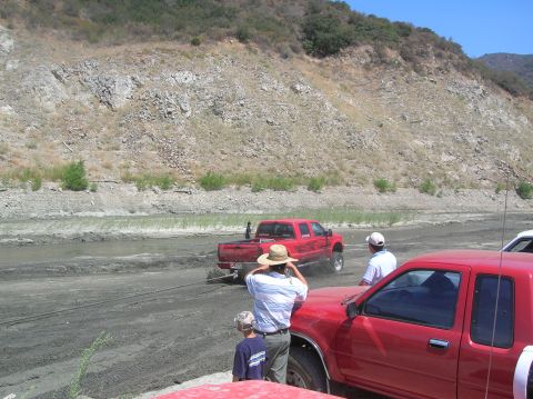 1985 Ford Bronco - Two red trucks are in a muddy area with a rocky hillside in the background.