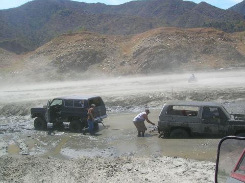 1985 Ford Bronco - Two SUVs are stuck in a muddy area, with people attempting to pull one out with a tow strap.