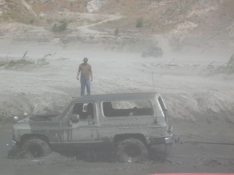 1985 Ford Bronco - A man stands on the roof of a mud-covered SUV stuck in a muddy terrain, with another vehicle in the distance.