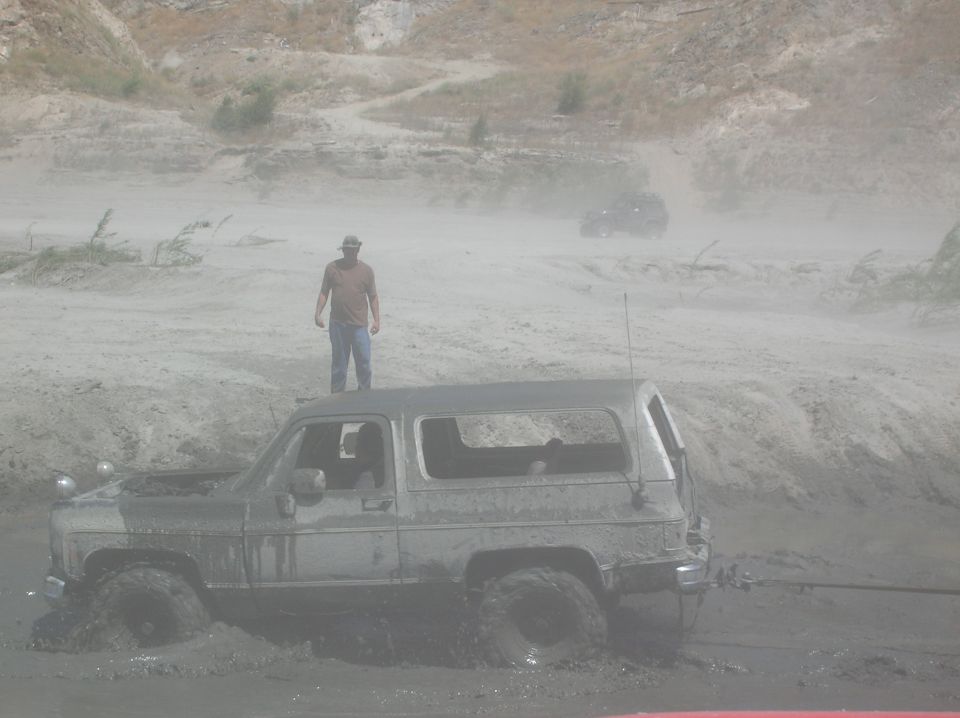 Muddy SUV stuck in mud, person standing on roof. Another SUV in background