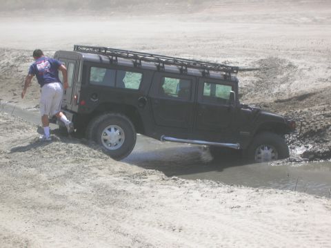 1985 Ford Bronco - A black Hummer H1 is partially submerged in mud as a man walks beside it.