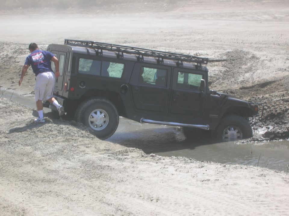 Black Hummer H1 stuck in mud with a man in a blue shirt and khaki shorts nearby