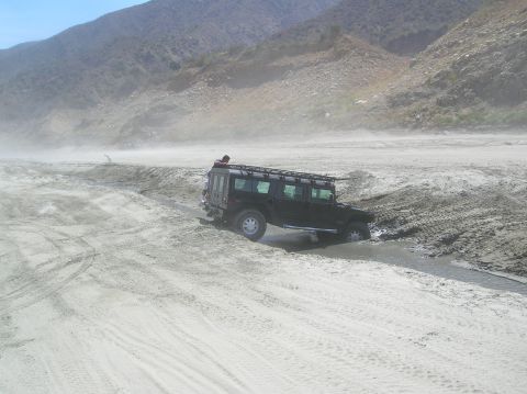 1985 Ford Bronco - A black SUV navigates a sandy, watery terrain, kicking up dust under a clear sky.