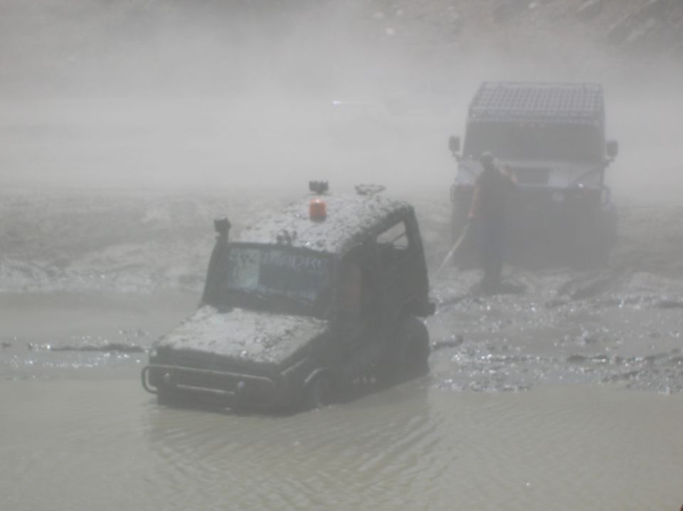 Two mud-covered off-road vehicles stuck in muddy water, one in the foreground with an orange light on top