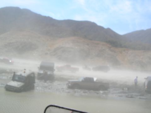 1985 Ford Bronco - Multiple off-road vehicles are seen traversing a challenging muddy and dusty terrain under a bright sky.