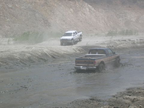 1985 Ford Bronco - Two pickup trucks navigate a muddy terrain, kicking up spray as they go.