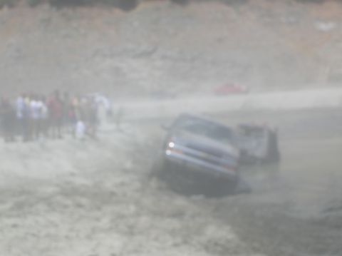 1985 Ford Bronco - A dark SUV is stuck in the mud, with a crowd of onlookers observing the scene. Another vehicle is partially visible in the background.