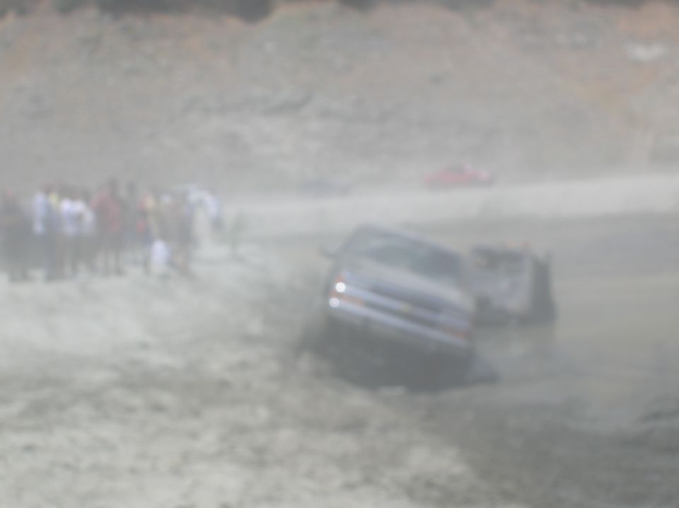 Dark SUV stuck in mud, crowd gathered, another vehicle partially visible