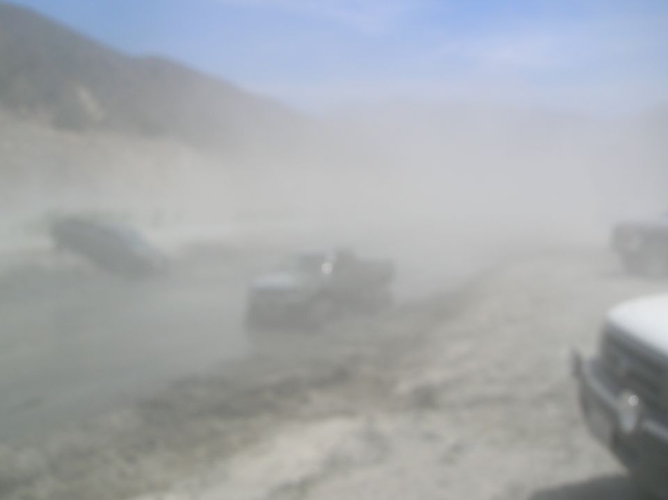 Several vehicles driving on a dirt road, kicking up a large dust cloud. Hills in background