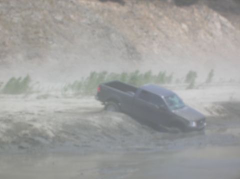 1985 Ford Bronco - A dark-colored pickup truck is stuck in muddy terrain, with vegetation visible in the background.