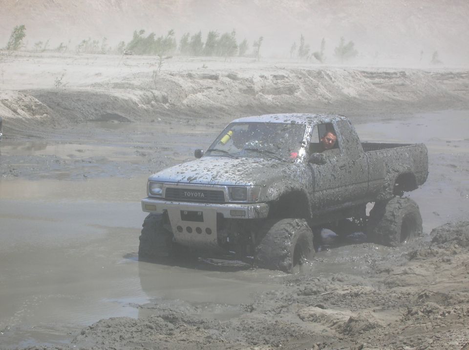 Mud-covered Toyota pickup truck with large tires driving through a muddy field. Driver is visible