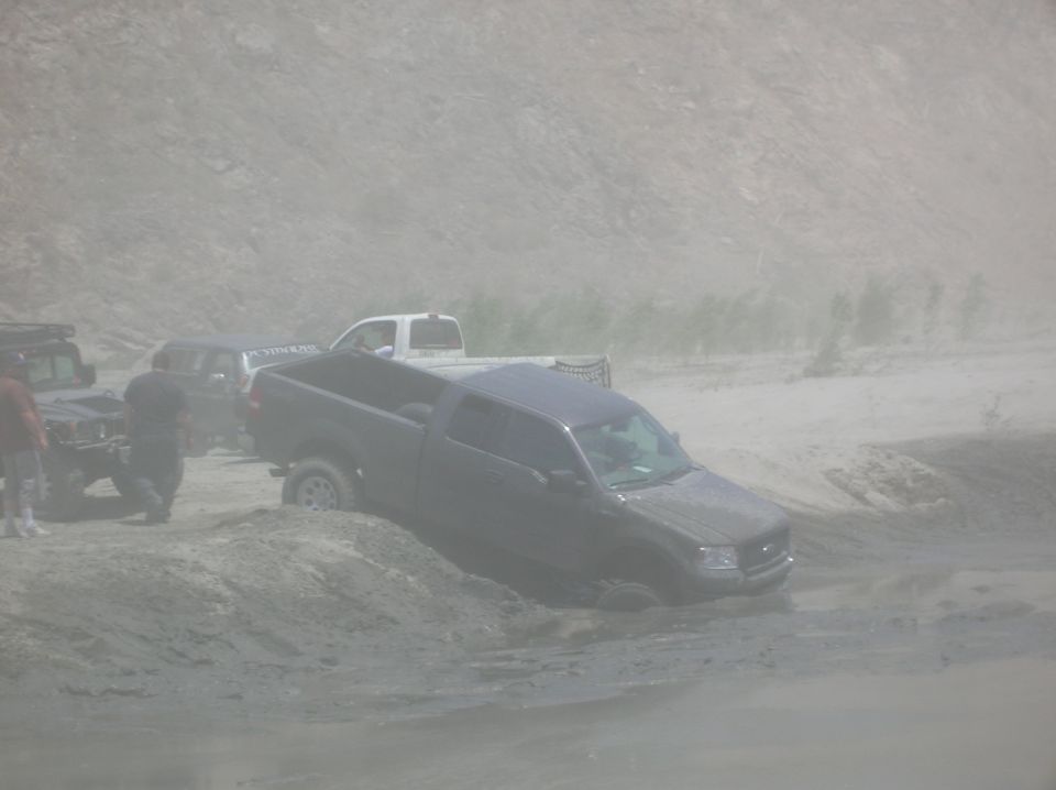 Several trucks stuck in sand dunes, one dark pickup truck is partially submerged
