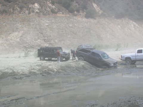 1985 Ford Bronco - Multiple trucks and SUVs are stuck in a muddy, dusty off-road area with a rocky incline in the background.