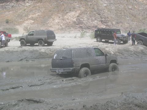 1985 Ford Bronco - Several vehicles navigate a muddy terrain, with one SUV deeply stuck in the mire.