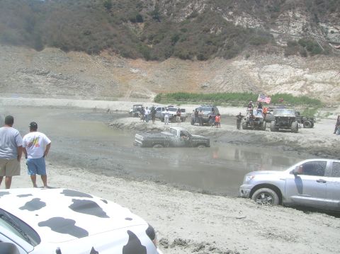 1985 Ford Bronco - Several off-road vehicles are gathered around a muddy pickup truck that is stuck in a mud pit.
