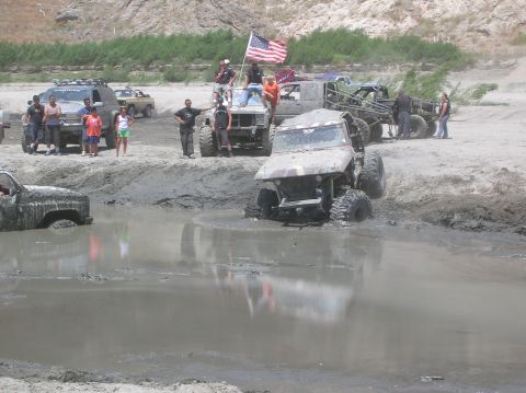 1985 Ford Bronco - Multiple off-road vehicles are stuck in a muddy area, with spectators gathered around and an American flag flying.