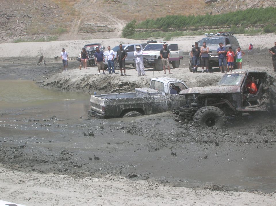 Two off-road trucks stuck deep in mud with people watching from the bank