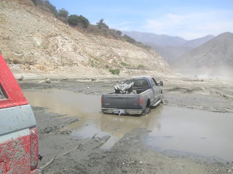 1985 Ford Bronco - A dark blue pickup truck, carrying an ATV, is stuck in thick mud in a dry riverbed, with another vehicle partially visible in the foregroun…