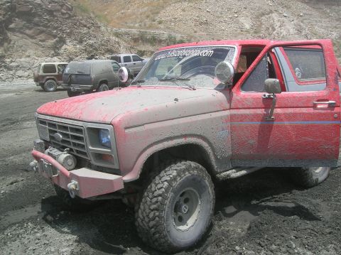 1985 Ford Bronco - Muddy 1985 Ford Bronco with website sticker on windshield, parked in a dirt lot with other vehicles.