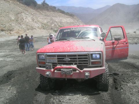 1985 Ford Bronco - This red Ford Bronco is stuck in the mud with its door open. People are standing around in the background.