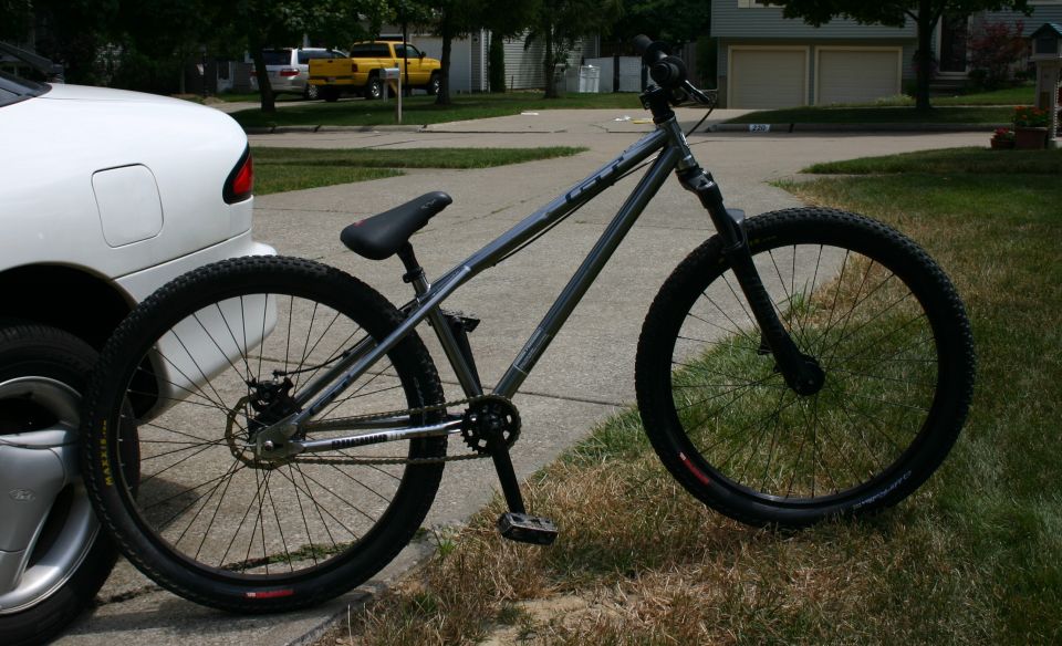 Dirt jump bicycle parked next to a white car. Bicycle has a silver frame and black seat and tires