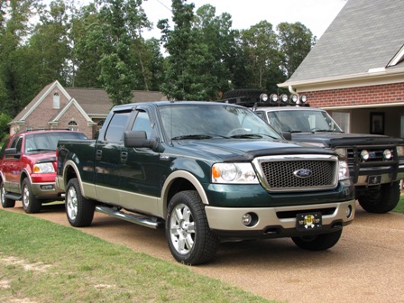 1991 Ford Bronco - Three Ford trucks, including a green F-150 and a red SUV, are parked in a residential driveway.