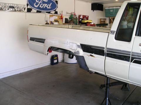 1986 Toyota Hilux - White Toyota pickup bed and cab sitting on jack stands in a garage with the frame removed from underneath, the bed floating beside a black…