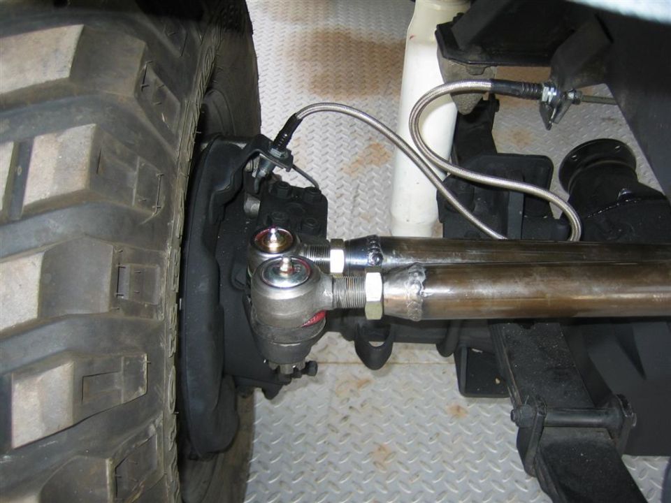 Heavy-duty tie-rod ends on a welded steel tie rod with braided brake line behind a mud tire