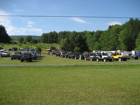 1991 Ford Bronco - A large gathering of off-road vehicles are parked in a grassy area, with a line of trees and a blue sky visible.
