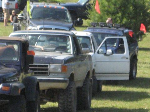 1991 Ford Bronco - A line of modified trucks and SUVs are parked on a grassy field.