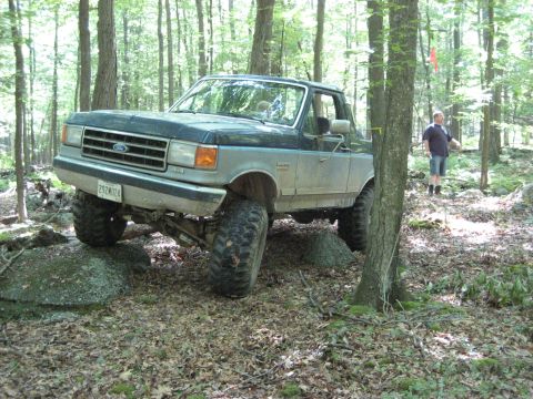 1991 Ford Bronco - A lifted Ford F-Series truck navigates rocky terrain in a wooded area, with a person standing in the background.