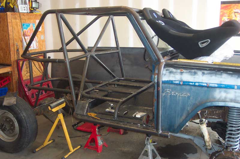 Side view of a blue Ford Bronco undergoing modification with a prominent roll cage and racing seats