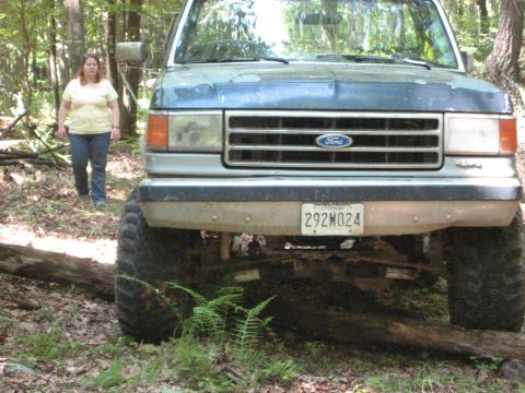 1991 Ford Bronco - A blue Ford Bronco is seen from the front in a forest, with a woman standing in the background.