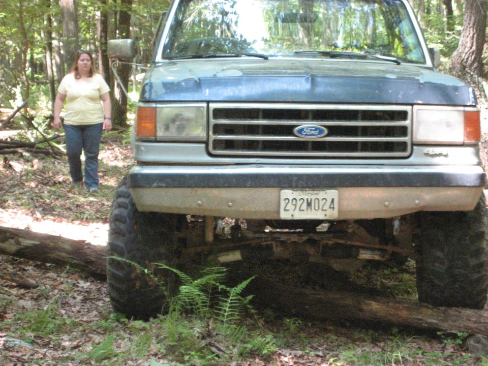 Blue Ford Bronco with a lifted suspension and large tires, parked in a wooded area. Front view