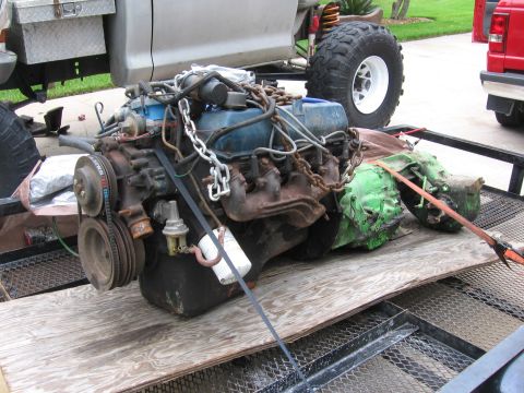 1974 Ford F-100 - A V8 engine and transmission are secured on a trailer, with a truck bed and a red truck visible in the background.