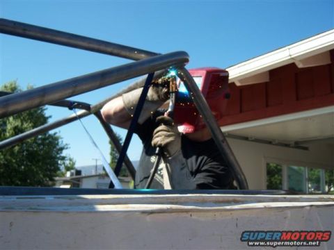 1996 Ford Bronco - Welder fabricates a roll cage, sparks fly against a blue sky and building.