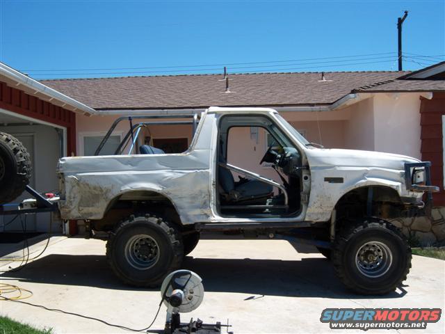 White Ford Bronco with roll cage, missing doors, and off-road tires, parked outside a house