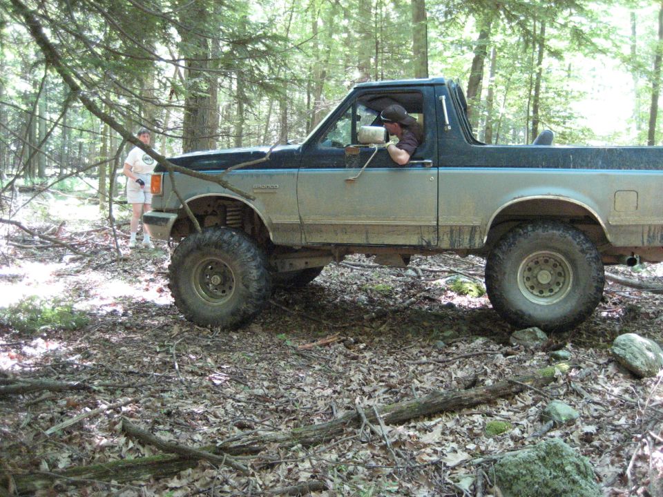 Ford Bronco with lifted suspension and large tires driving through a wooded area. A person stands in the background