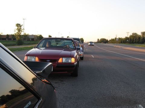 1989 Ford Mustang - A dark red Fox-body Ford Mustang LX with its occupants inside idles on a roadside shoulder at sunset, shot from behind a black Fox-body Mus…