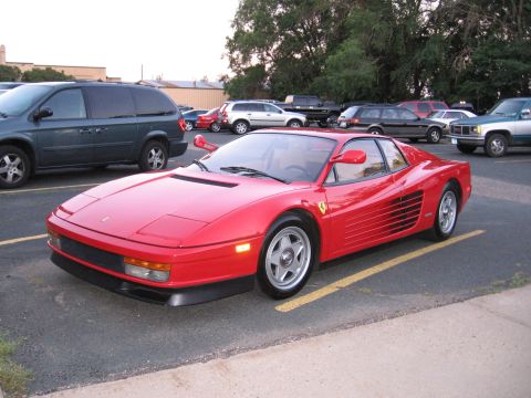 1989 Ford Mustang - A red Ferrari Testarossa is parked in a lot, with other vehicles visible in the background.