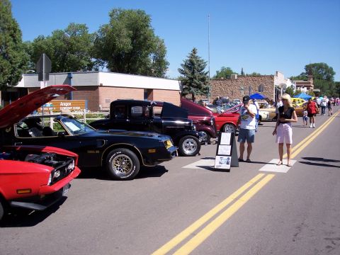 1972 Ford Ranchero - A line of classic cars are displayed on a street, including a black 1977 Pontiac Trans Am with its hood up.