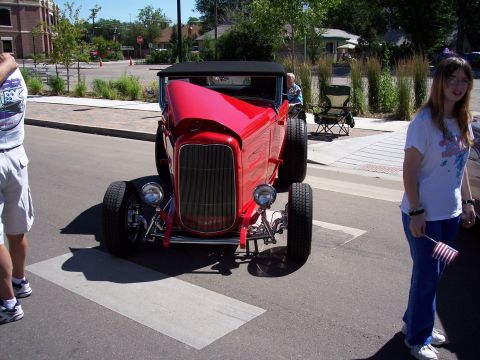 1972 Ford Ranchero - A bright red hot rod is parked on a street, with onlookers and a woman holding a small American flag.