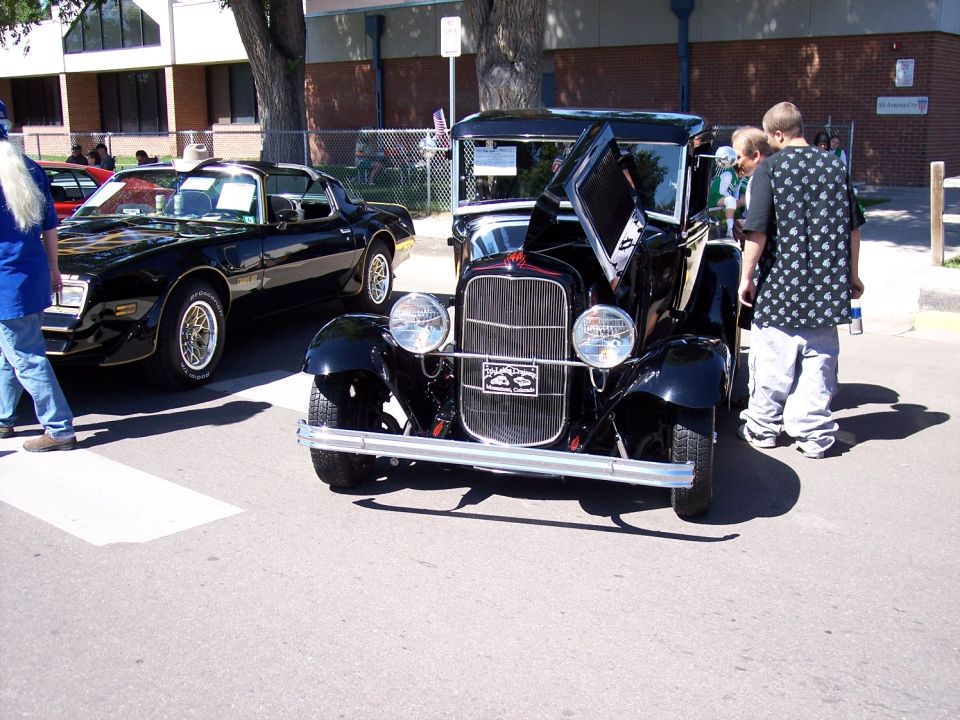 Black vintage car with hood open, chrome bumper, and grille. A black 1980s Pontiac Firebird is parked to the left