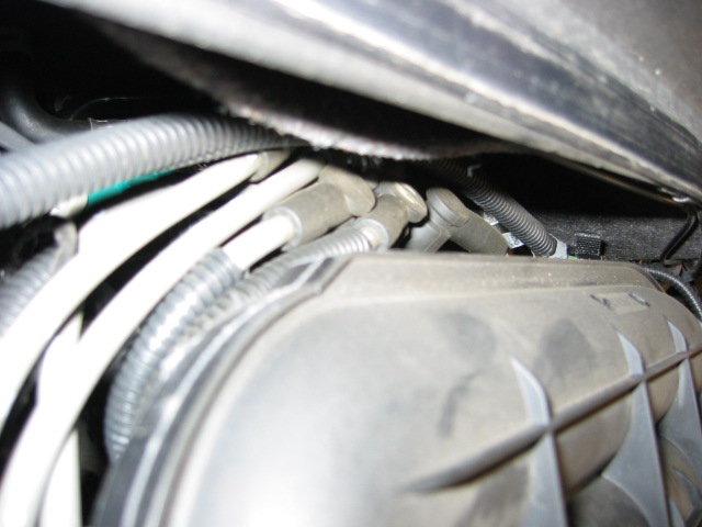 Blurry under-hood close-up of a ribbed black housing, wiring looms and a threaded stud