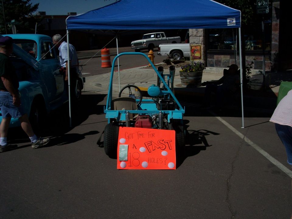 Blue go-kart with orange sign that says "GOT TIME FOR A FAST 18 HOLES?" parked next to a light blue pickup truck