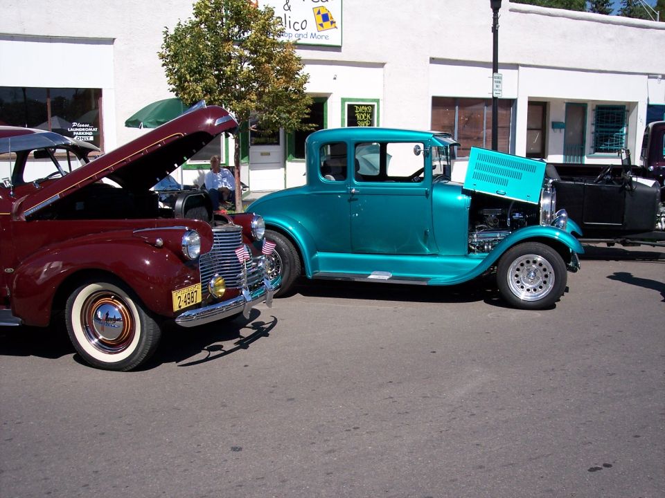 Two classic cars parked on a street, one maroon with hood open, the other teal with hood open showing engine
