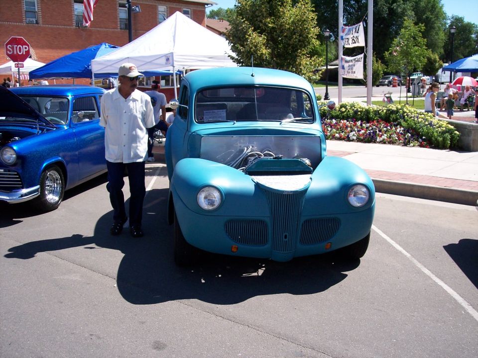 teal hot rod truck with hood open, man standing next to it, blue car to the left